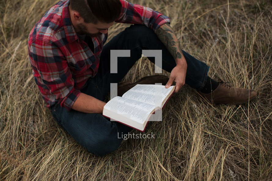 man in a plaid shirt sitting on the ground reading a Bible 