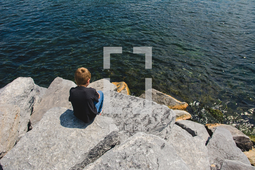 a little boy sitting on a rock along a shore 