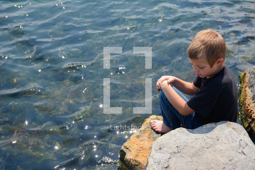 a boy sitting on a rock on a lake shore 