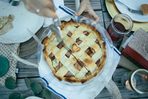 Hands cutting an apple pie on a picnic table outside.
