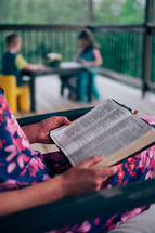 a mother reading a Bible while her kids color on a porch 