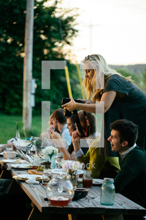a woman taking a picture with a camera as a family eats dinner together at a table outdoors 