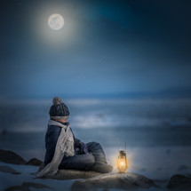 a boy sitting on a shore in winter at night next to a lantern 