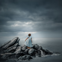 a girl in a dress standing on rocks along a shore 