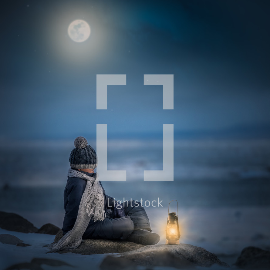 a boy sitting on a shore in winter at night next to a lantern 