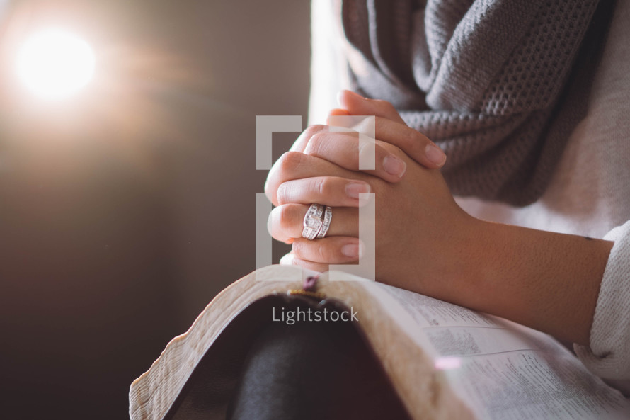A woman sits with hands clasped on an open Bible.