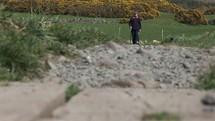 Man walking with his dog across a meadow and stony area.  Yellow brush in the background