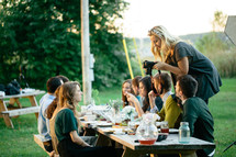 a woman taking a picture with a camera as a family eats dinner together at a table outdoors 