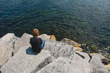 a little boy sitting on a rock along a shore 