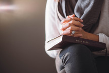 A woman sits with hands clasped on a Bible.