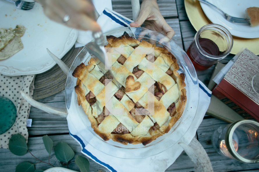 Hands cutting an apple pie on a picnic table outside.