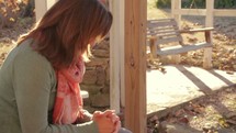 Woman praying while sitting on a bench on the porch outside.