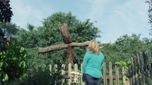 Woman praying at a wooden cross in a garden.