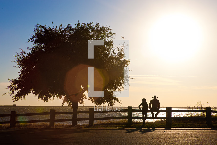 A couple sits on a fence at the edge of a lake at sunset