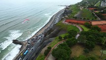 Aerial shot drone tracks paraglider as it flies over traffic heavy coastal highway with waves crashing against rocky beach and high green gardens on cliffs