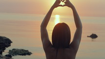 Young woman shapes heart with hands over the sunset by the ocean