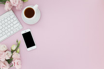 Computer keyboard, cellphone, cup of coffee and pink flowers on a pink background.
