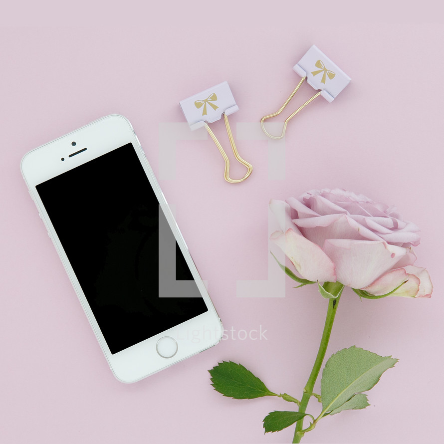 A cellphone, gold clips and a pink rose on a pink background.