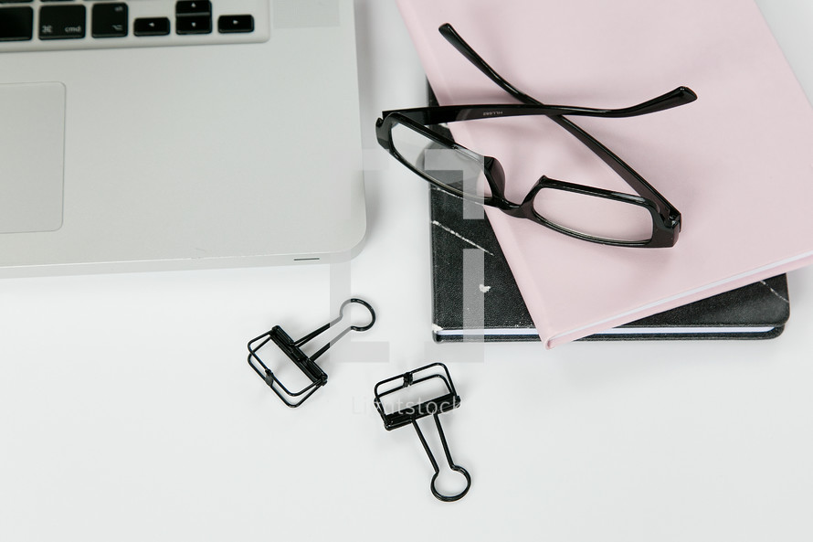 reading glasses, clips, journals, and laptop on a white desk 