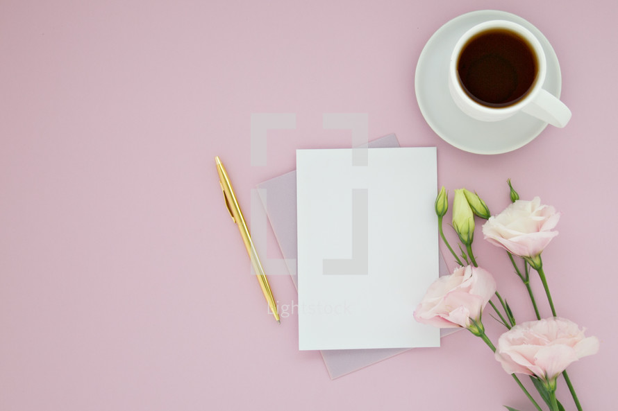 A blank notecard, gold pen, cup of coffee and pink flowers on a pink background.