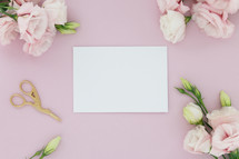A blank white card on a pink background surrounded  by flowers and scissors.