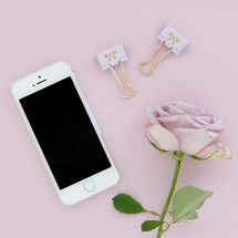 A cellphone, gold clips and a pink rose on a pink background.