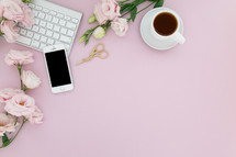 A computer keyboard, cell phone, cup of coffee and pink flowers on a pink background.