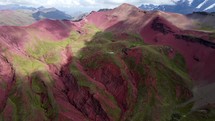 Aerial shot drone flies forward toward lake in bright red mountains with green vegetation and snow capped mountains in the distance