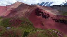 Aerial shot drone flies toward rock formation on bright red mountains with green vegetation and snow capped mountains in the distance