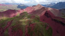 Aerial shot drone hovers over bright red mountains with green vegetation and snow capped mountains in the distance