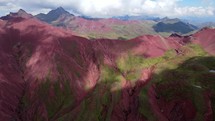 Aerial shot drone orbits to the left in front of bright red mountains with green vegetation and snow capped mountains in the distance